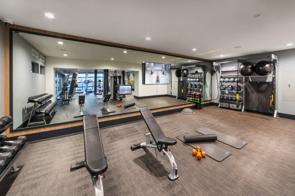 Fitness center room with flat screen TV, mirrored walls, benches, free weights, yoga mats and medicine balls.