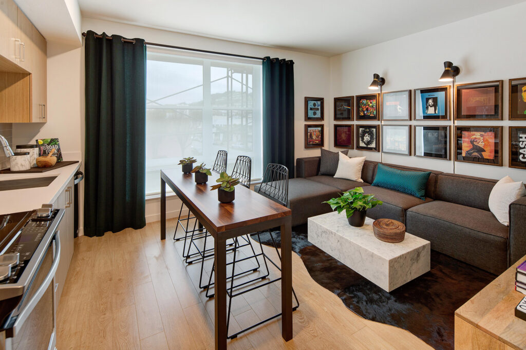 Open concept living room/kitchen with wood-style floor, stainless steel appliances, large window, kitchen island with seating and sectional couch.
