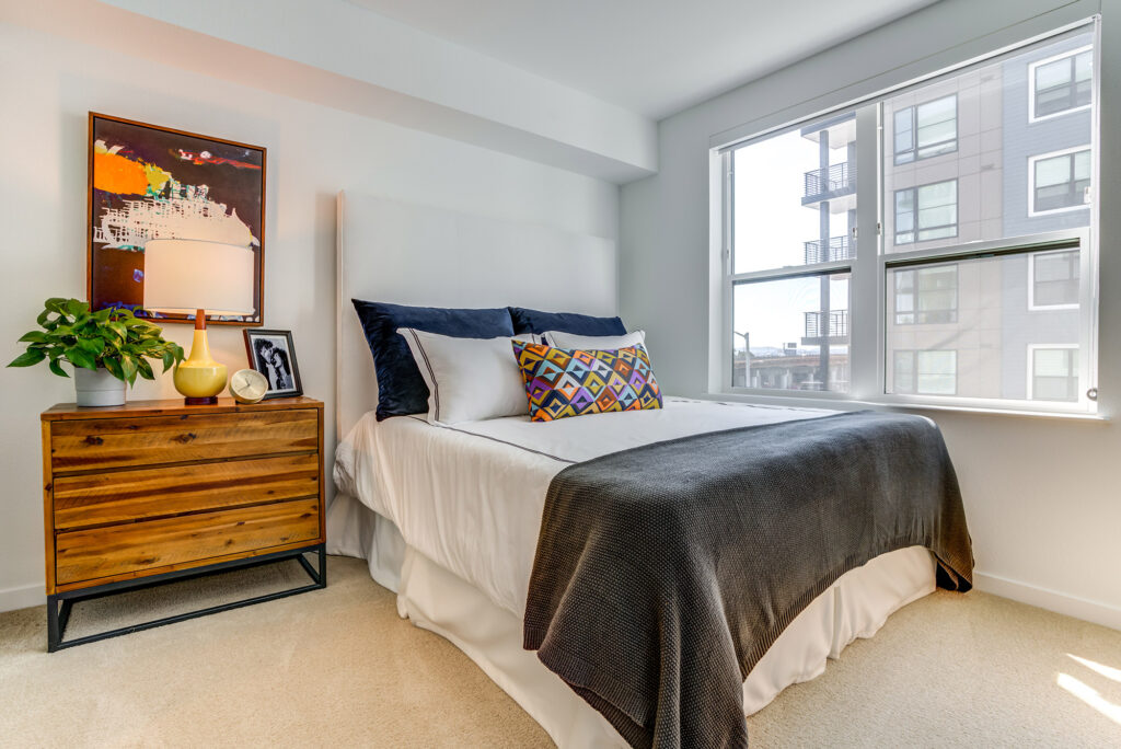 Bedroom with plush carpeting, large window for natural light, queen-sized bed and nightstand.