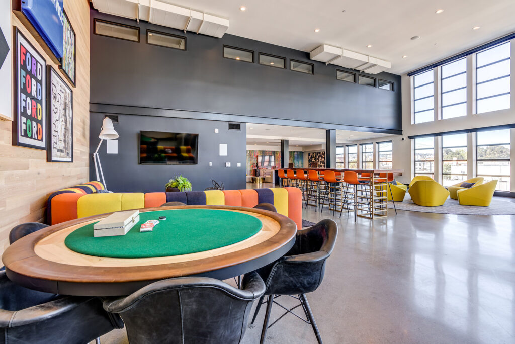 Clubroom with large family-style high-top seating, poker table, retro artwork, and large windows.
